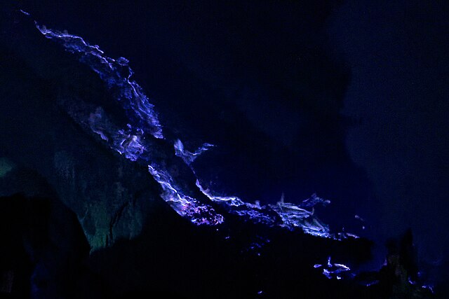 Blue flames flowing across the slope of a dark volcanic crater.