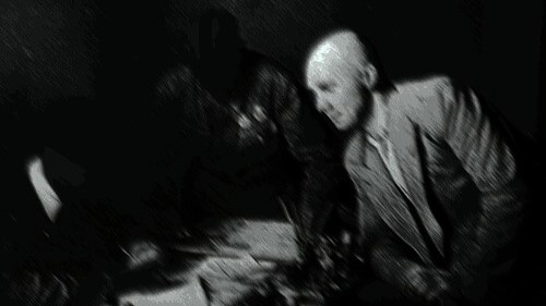 Unmasked figure leaning on a table with two masked figures — one seated and writing, the other standing and leaning on the side.