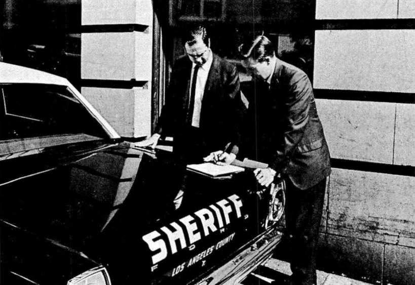 Black-and-white image of two obscure figures beside a car.