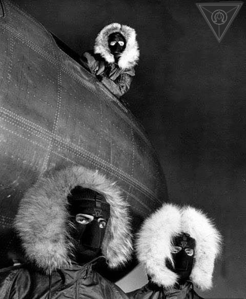 Two obscured figures standing beside an airship; one is lying on top, all facing the camera.